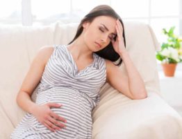Struggling with morning sickness. Depressed pregnant woman holding hand on head and keeping eyes closed while sitting on a couch
